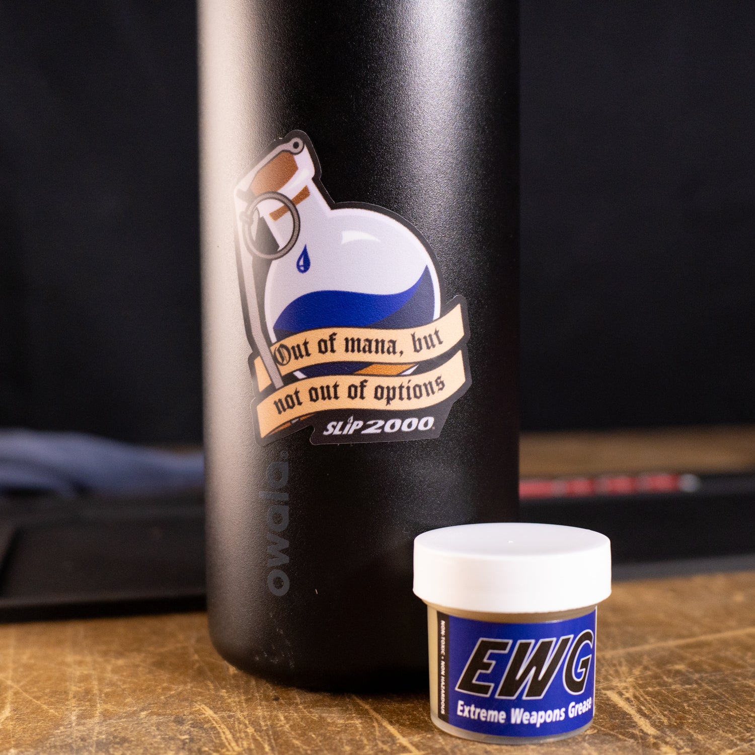Sticker of a blue potion bottle with a grenade spoon saying "out of mana, but not out of options" on a black bottle with a tub of Slip 2000 EWG next to it.
